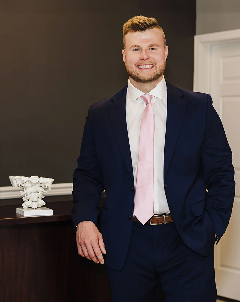 Dr. Robert Bender wearing a suit and pink tie, standing in front of a desk.